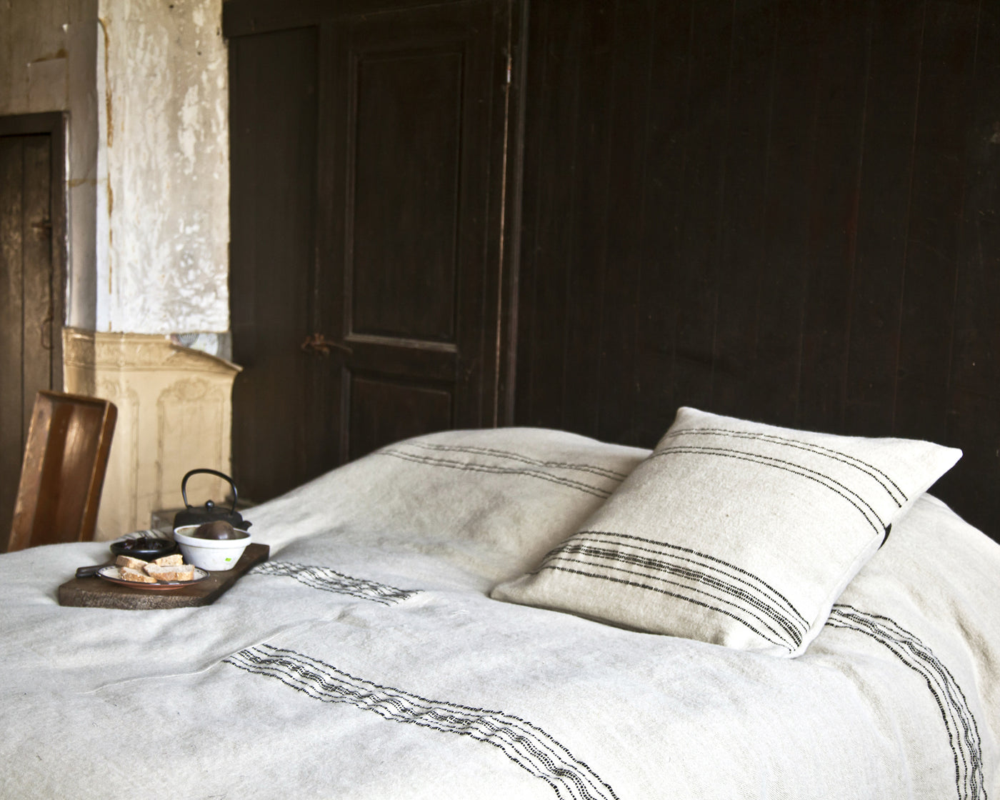 A neatly made bed with a white blanket featuring black stripes, a matching pillow, and a tray with tea and snacks placed on the blanket in a rustic room.