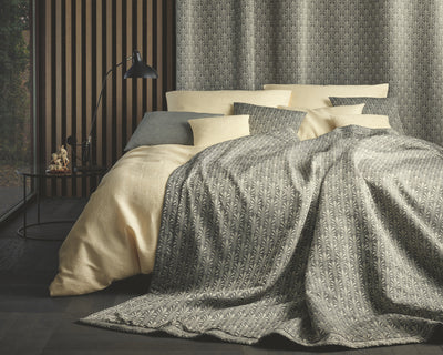 A neatly made bed with patterned grey and beige bedding, multiple pillows, a black floor lamp, and wood-paneled walls beside a large window.
