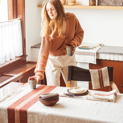 Woman setting a table with a Libeco ivory linen tablecloth and rust-striped table runner.