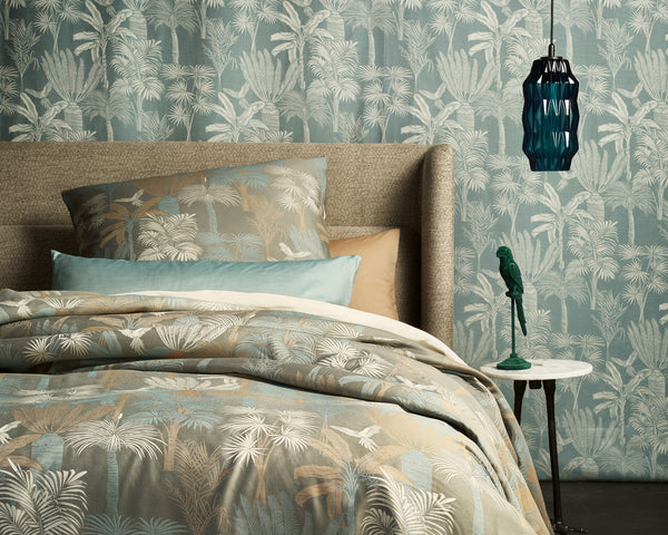 A bed with patterned bedding and pillows stands against a wall with matching wallpaper. A blue pendant light and a small side table with a green bird figurine are beside the bed.