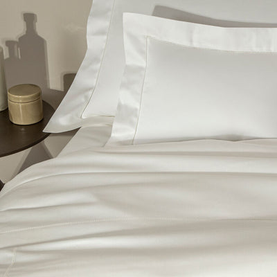 A neatly made bed with white sheets and pillowcases, next to a dark round side table holding a gold canister and a bottle.