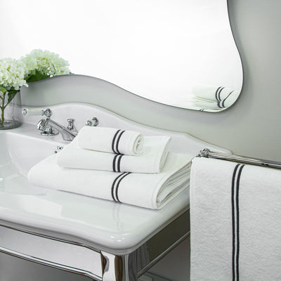 A stack of white towels with black stripes is arranged on a white bathroom sink beside a faucet, with flowers and a mirror in the background.