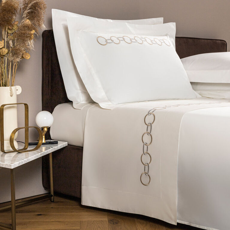 A neatly made bed with white sheets and pillowcases featuring a linked ring embroidery pattern, next to a bedside table with a lamp, vase, and dried plants.
