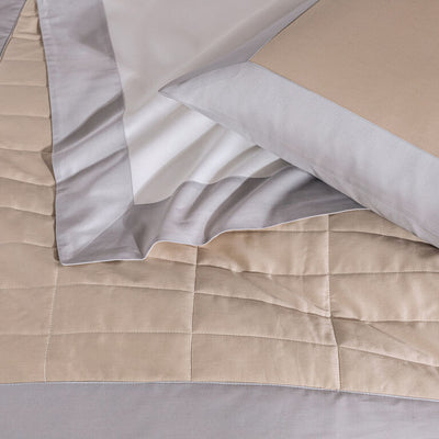 A neatly made bed with a beige and gray quilted comforter, matching pillow, and white sheet with a gray border.