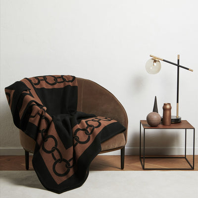 A brown chair with a black and brown patterned blanket draped over it sits next to a small table holding a lamp and decorative objects against a white wall.