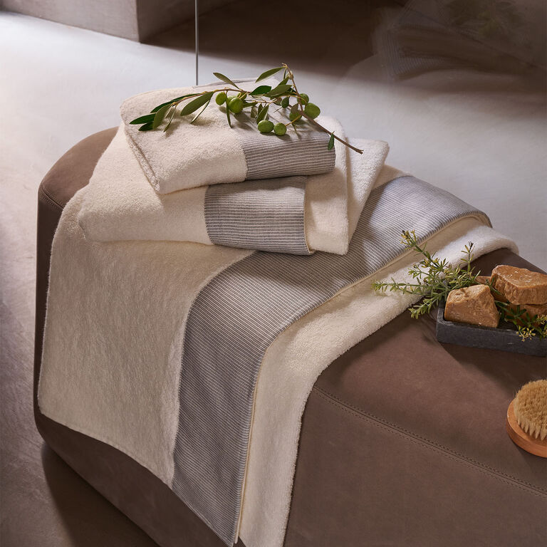 Stack of folded beige and gray striped towels with a sprig of greenery, placed on a brown ottoman next to soap bars and a wooden brush.