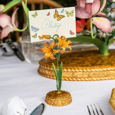 Orange lily place card holder on gold base holding butterfly-printed place card on white linen table.