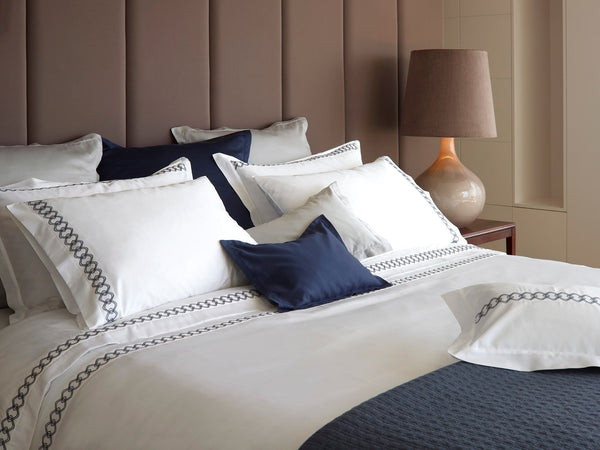 A neatly made bed with white and navy bedding, decorative pillows, and a brown upholstered headboard next to a brown lamp on a wooden nightstand.