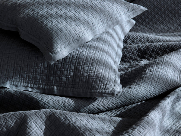 Two pillows and a crumpled quilted bedspread with a subtle grid pattern, shown in soft, angled light with visible shadows.