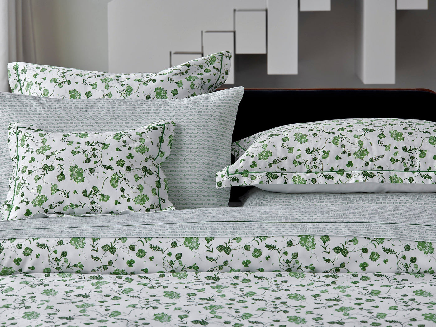 Bed with patterned white and green floral bedding, including pillows and sheets, set against a modern white and gray geometric background.