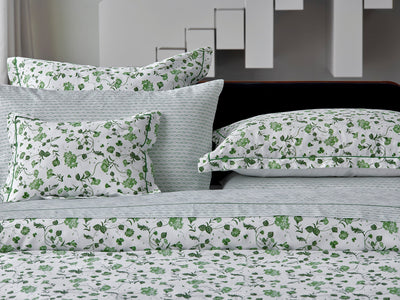 Bed with patterned white and green floral bedding, including pillows and sheets, set against a modern white and gray geometric background.