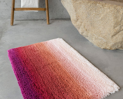 A rectangular rug with an ombre design in shades of pink, red, and white sits on a gray concrete floor near a wooden stool and a beige stone surface.