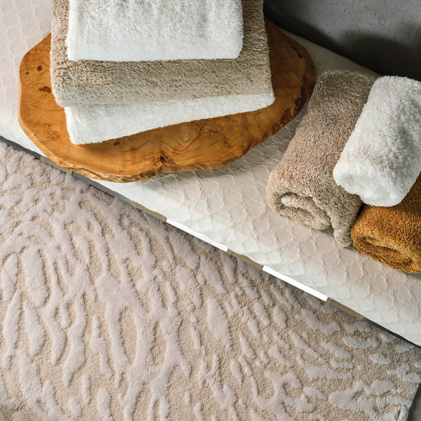 Folded white and beige towels are stacked on a wooden tray, placed on a textured surface next to a beige patterned rug.