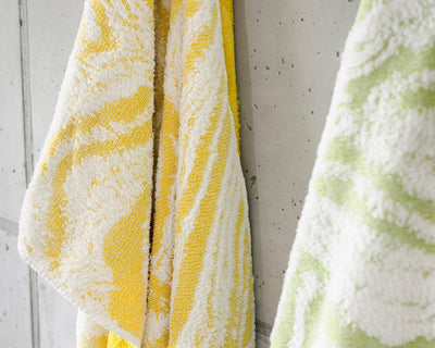 Two patterned towels, one yellow and white and the other green and white, hang side by side on a light gray tiled wall.
