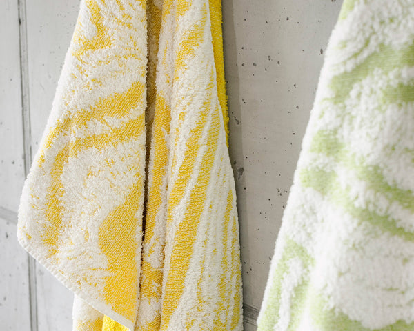 Two patterned towels, one yellow and white and the other green and white, hang side by side on a light gray tiled wall.
