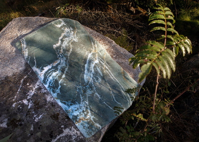 A slab of greenish blue rock with white streaks rests on a larger gray stone, surrounded by plants and natural outdoor lighting.