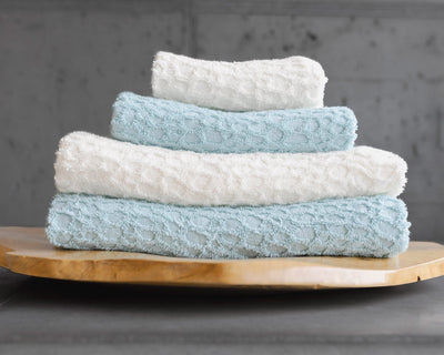 Four folded towels, two white and two light blue, stacked in alternating colors on a wooden tray against a gray background.
