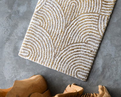 A rectangular rug with a textured, curved line pattern in white and gold on a gray concrete floor, next to the edge of a wooden chair.