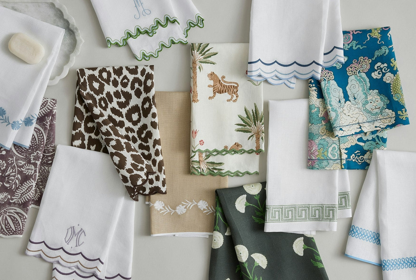 A variety of folded patterned and embroidered hand towels are arranged on a flat surface, featuring designs like animals, florals, and scalloped edges.