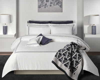 A neatly made bed with white bedding, navy accents, and geometric-patterned blanket. Two lamps sit on side shelves, and a modern abstract artwork hangs above the headboard.