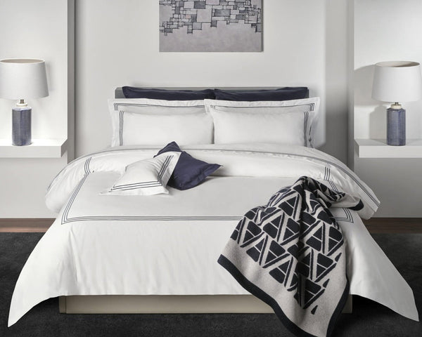 A neatly made bed with white bedding, navy accents, and geometric-patterned blanket. Two lamps sit on side shelves, and a modern abstract artwork hangs above the headboard.