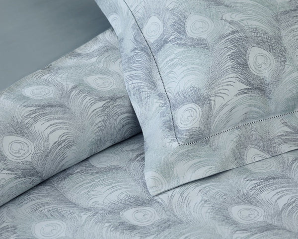 Close-up of bedding with a light gray and white peacock feather pattern, showing a pillow and part of a sheet with detailed stitching.