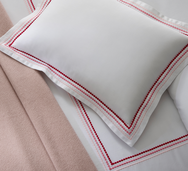 White pillow and sheet with red embroidered borders on a bed, next to a light pink blanket.