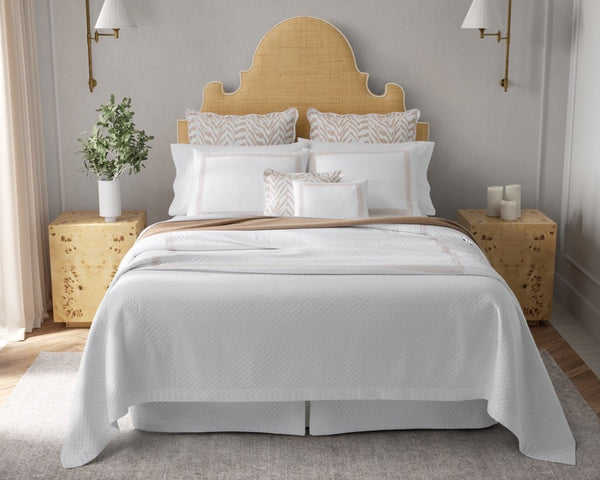 A neatly made bed with white bedding, beige headboard, decorative pillows, two wooden nightstands, a plant, candles, and wall-mounted lamps in a bright bedroom.