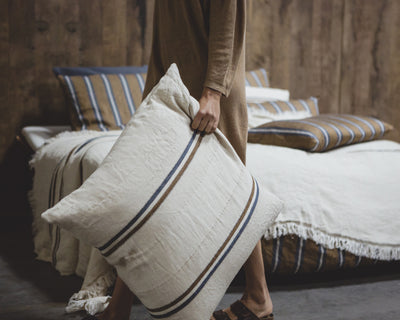 Person holding a large cream-colored striped pillow in a bedroom with a wooden wall, a bed, and striped bedding in the background.