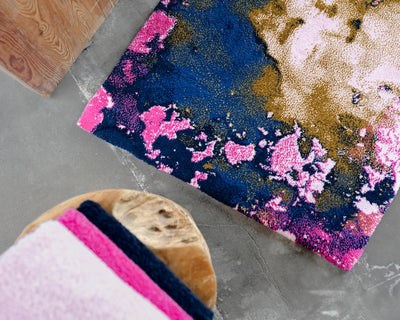 A colorful rug with navy blue, pink, and gold abstract patterns on a concrete floor next to a wooden table with folded towels in pink, navy, and white.