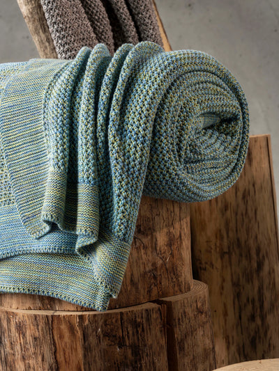 A close-up of a folded gray knit blanket draped over a bed with a geometric patterned white and gray sheet.