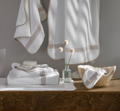 White towels with beige decorative trim are neatly arranged on a wooden surface, with a bar of soap, a small glass vase with flowers, and a matching bowl.