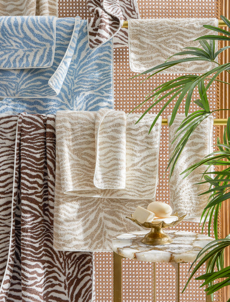 Zebra-patterned towels in blue, beige, and brown hang on a gold rack next to a marble table with a brass dish holding bars of soap and a green plant in the foreground.