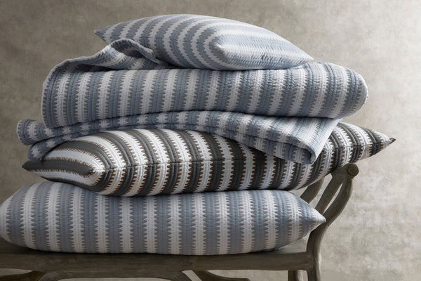 A stack of patterned pillows and folded blankets in shades of blue, gray, and white rests on a wooden chair against a neutral background.