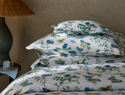 A neatly stacked bed with white bedding and pillows featuring a blue and yellow floral and butterfly print, next to a lamp and a striped mug on a side table.