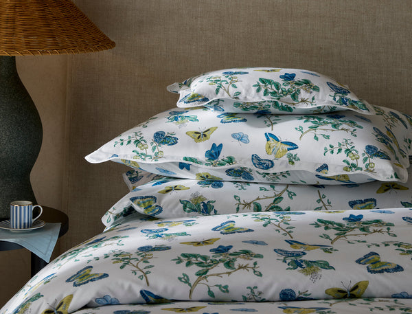 A neatly stacked bed with white bedding and pillows featuring a blue and yellow floral and butterfly print, next to a lamp and a striped mug on a side table.