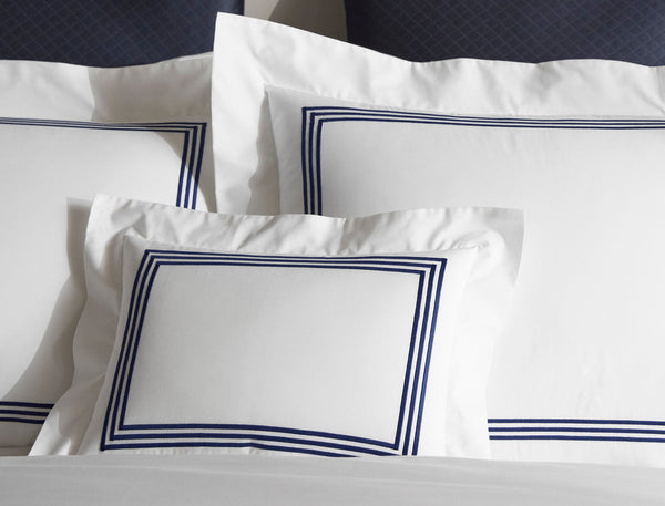 Three white pillows with navy blue double-line borders are arranged on a bed with matching white sheets and two dark blue pillows in the background.
