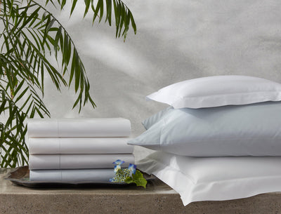 A stack of folded bed sheets and a pile of pillows in white and light gray, arranged on a concrete surface with a green plant in the background.