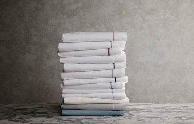 A stack of folded white bed sheets with colored trim sits on a marble surface against a gray textured background.