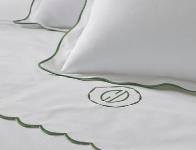 Close-up of white bedding with green scalloped edges and an embroidered circular monogram, featuring two pillows and part of a sheet or duvet.