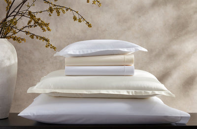 A neatly stacked set of white and cream-colored pillows and folded bed sheets sits beside a large vase with yellow flowering branches.