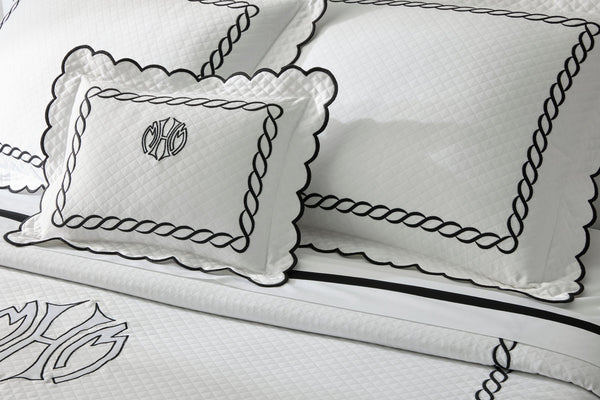 White bedding set with quilted texture, black embroidered borders, and monogram detail on pillows and duvet cover.