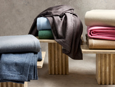 Three wooden benches display folded wool blankets in various colors, including blue, green, brown, beige, and pink, against a neutral background.
