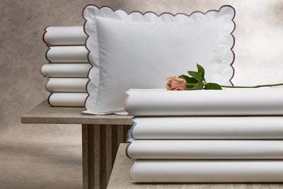 A single pink rose on a stack of folded white sheets with colored trim, next to a scalloped-edge pillow on a stone surface.