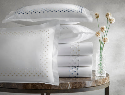 Stacked white pillowcases and sheets with embroidered dots in beige and blue, next to a glass vase with dried flowers on a round marble table.