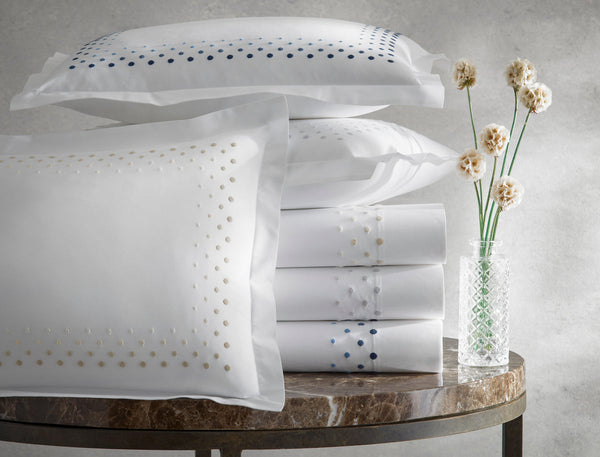 Stacked white pillowcases and sheets with embroidered dots in beige and blue, next to a glass vase with dried flowers on a round marble table.