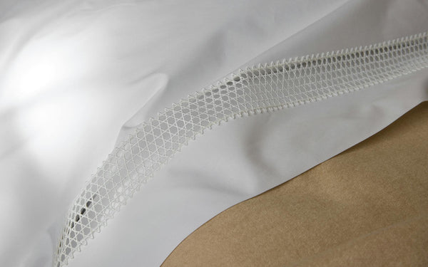 White fabric with a decorative net-like lace trim is draped over a tan surface in soft lighting.