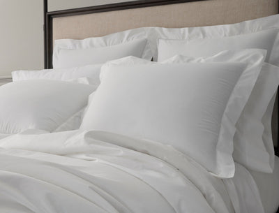 A neatly made bed with multiple white pillows and white bed sheets against a beige upholstered headboard.