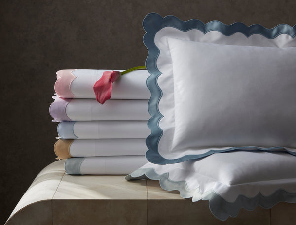 A stack of folded white bedsheets with colored scalloped edges sits next to two matching pillows; a pink calla lily rests on top of the stack.