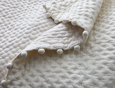 A close-up of a textured, cream-colored quilt with raised stitching and a border of small fabric pom-poms.